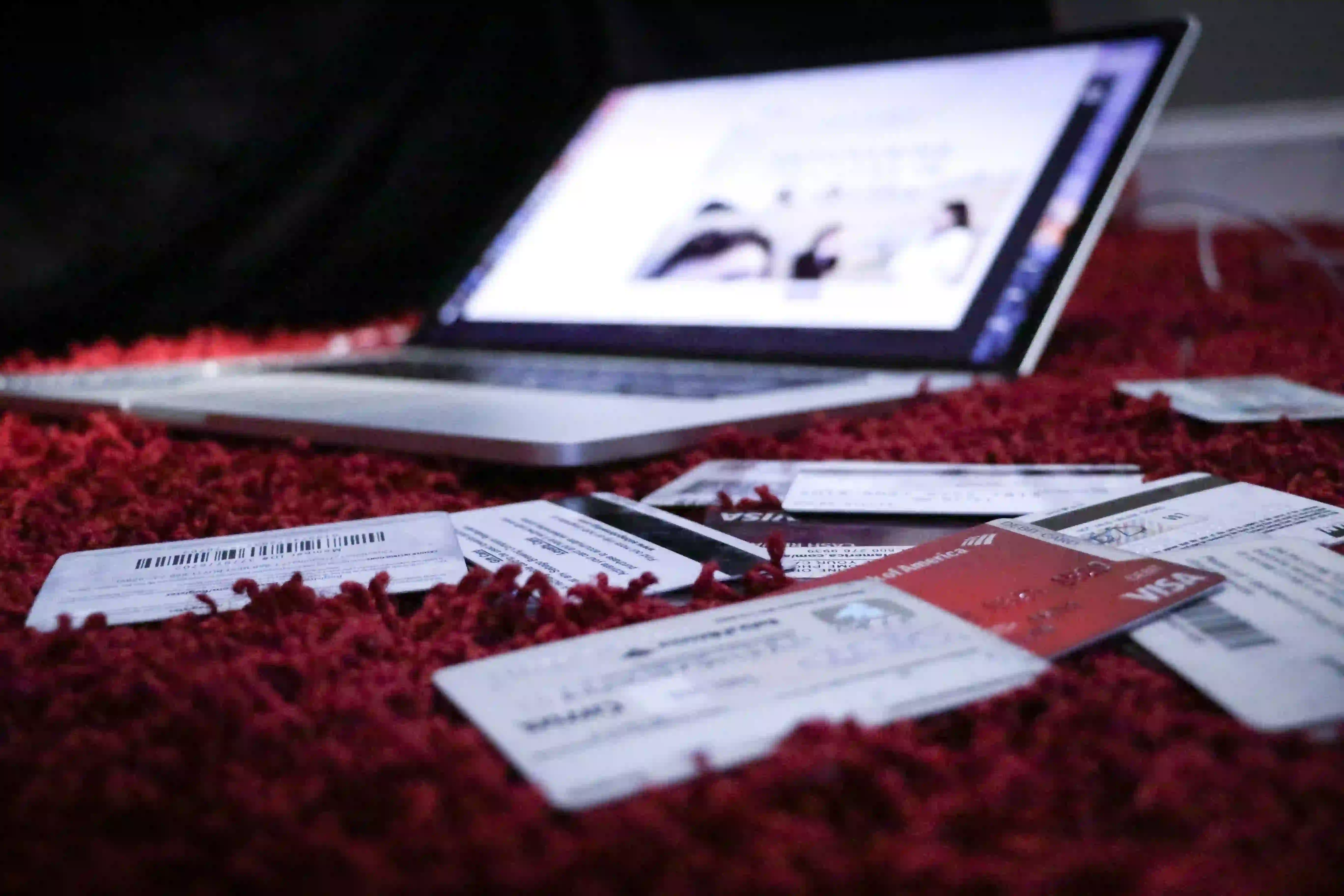 multiple credit cards scattered on a red carpet near a powered on laptop